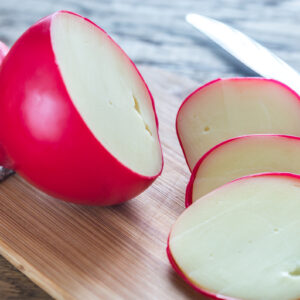 Cheese, wrapped in a red wax coating, is sliced and displayed on a cutting board.